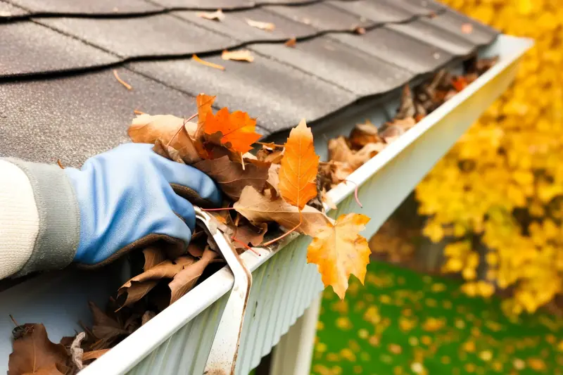 A gloved hand lifting wet autumn leaves out of a white house gutter at the edge of a roof