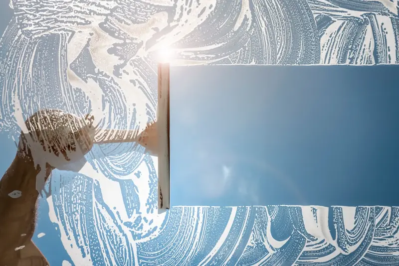 A squeegee pulling a clean strip through soapy water on a window, with blue sky behind the glass