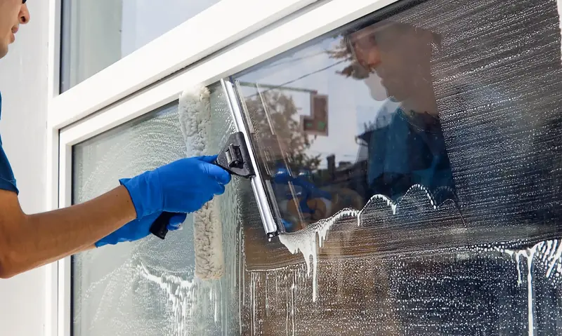 A window cleaner in blue gloves squeegeeing soapy water down a large ground-floor window