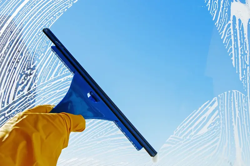 A blue squeegee in a yellow-gloved hand wiping soapy water off a window against a clear blue sky