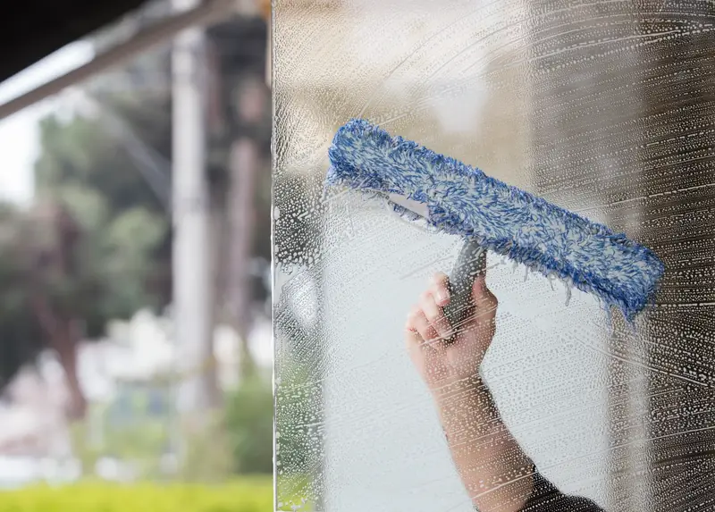 A blue microfibre window washer being drawn across soapy glass, with trees blurred in the background